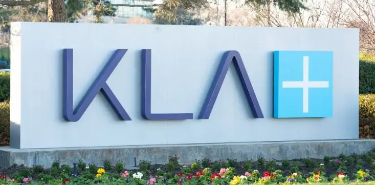 KLA Corporation logo displayed on a white monument sign, featuring purple letters and a blue square with a white plus symbol, surrounded by colorful flowers