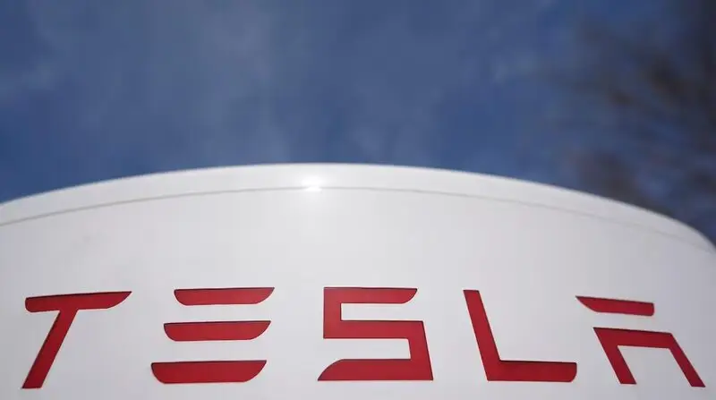 Close-up of red Tesla logo on a white curved surface against a blue sky background